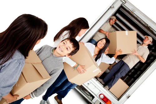 A group of young adults and children are engaged in packing and loading household items for a house move in Finsbury Park. They are standing in an outdoor residential setting, with some holding large cardboard boxes filled with various belongings. The children and adults are dressed casually, and their expressions appear focused and cooperative. In the background, a large white removals van is positioned with its rear doors open, revealing an interior filled with more boxes and household items, including a small box on the floor near the entrance. The scene is well-lit with natural daylight, illustrating a typical house removal activity with a focus on careful handling of belongings. The setting reflects a suburban residential area in or near Finsbury Park, with the scene emphasizing professional house removal services by Finsbury Park Man with Van, showcasing teamwork and organization during the packing and loading process.